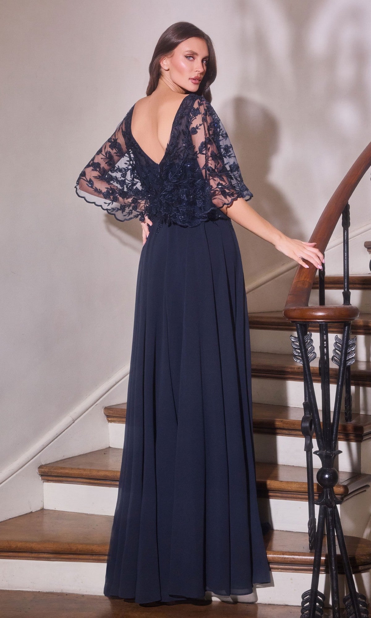 A woman in the Ladivine CL10 long formal dress, featuring lace sleeves, a deep V-back, and a lace-embroidered bodice, stands on a wooden staircase with one hand on the banister, showcasing its elegant flowing silhouette.